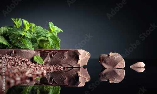 Pieces of dark bitter chocolate and chocolate chips with mint on a black reflective background.