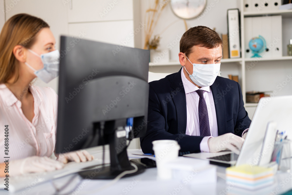 Fototapeta premium Portrait of busy entrepreneur in medical face mask and latex gloves working with female coworker in office. Concept of precautions and social distancing in coronavirus pandemic..