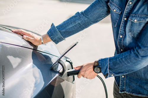 Close up cropped image of hands of man in jeans shirt and his electric car at charging station with the power cable supply plugged in. Electric cars, future car concept