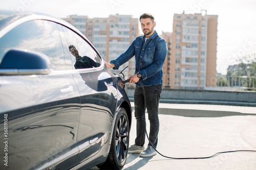 Full length portrait of young handsome bearded man in casual wear, standing at the charging station and holding a plug of the charger for an electric car. Eco electric car concept © sofiko14