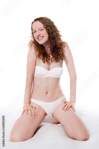 Beautiful laughing woman wearing panties and bra, she is kneeling on a bed in front of white background