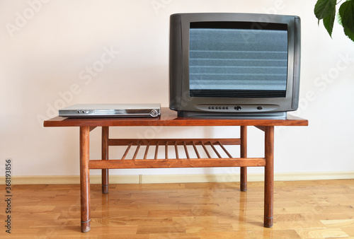 A DVD player and an outdated black TV with clutter on the screen sit on a vintage table in a 1990s apartment block. photo