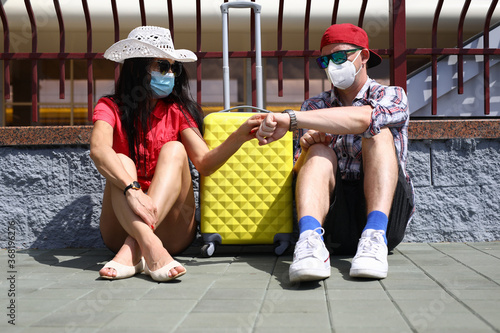 A guy and a girl in medical masks are sitting on the street. Young people with a suitcase travel during quarantine