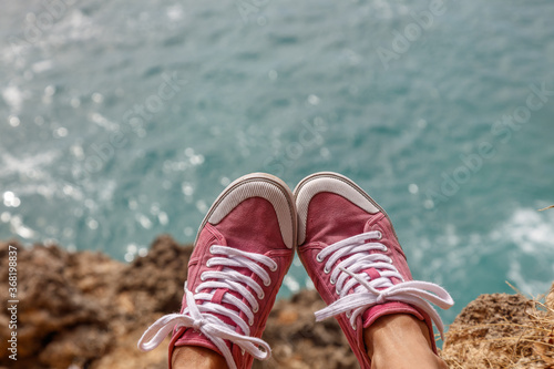 Girl in pink sneakers sitting on a cliff above the ocean. No face. Balangan Beach, Bukit, Bali, Indonesia.
