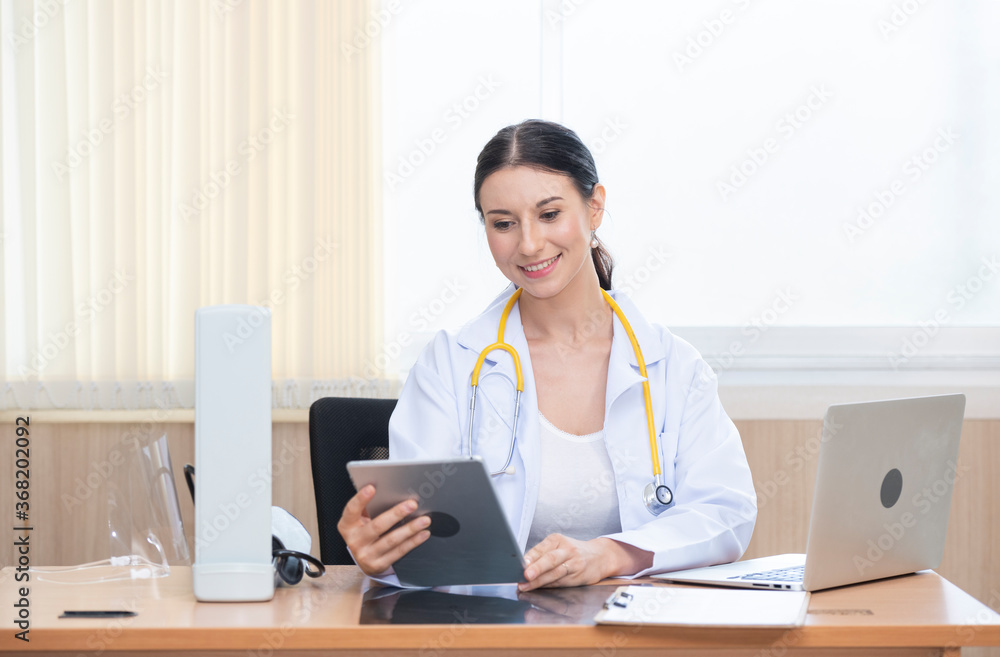 Female doctor with stethoscope sitting use tablet and computer laptop in office hospital. Confident woman doctor sitting at office desk, health care and prevention technology network concept Female doctor with stethoscope sitting use tablet and computer laptop in office hospital. Confident woman doctor sitting at office desk, health care and prevention technology network concept
