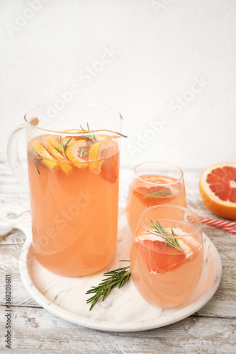 A glass jug and two glasses full of lemonade with grapefruit slices and sprigs of rosemary stand on a tray on a light wooden table. Nearby lies a half of grapefruit and a sprig of rosemary. Vertically