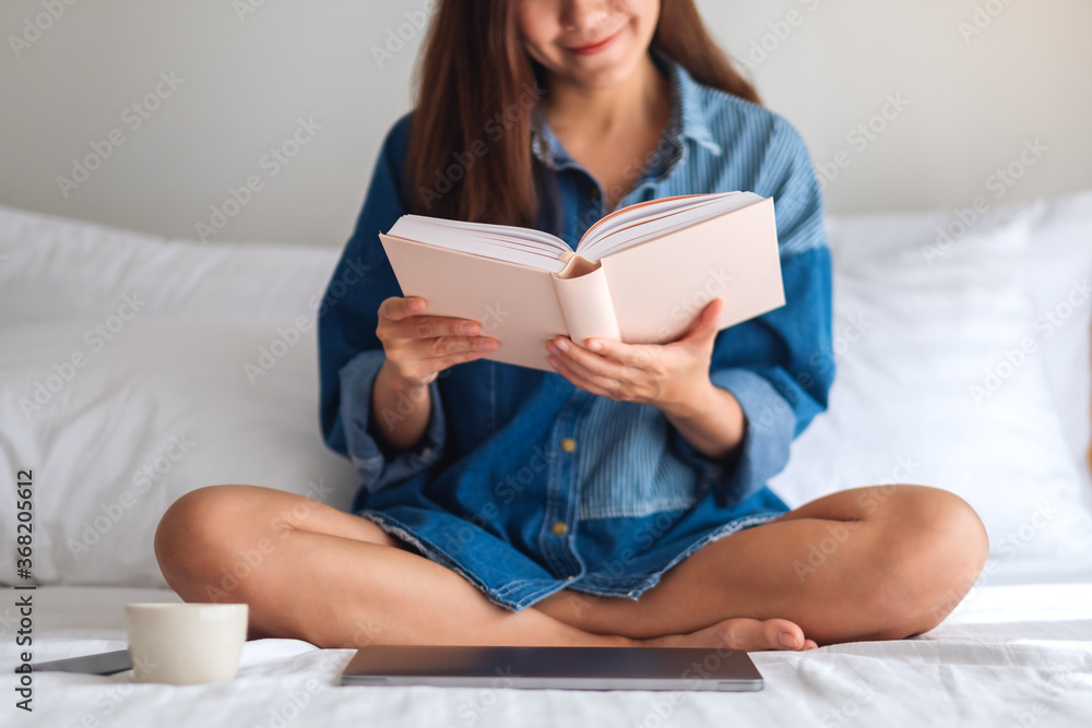 A beautiful young woman reading book while sitting on a white cozy bed at home A beautiful young woman reading book while sitting on a white cozy bed at home