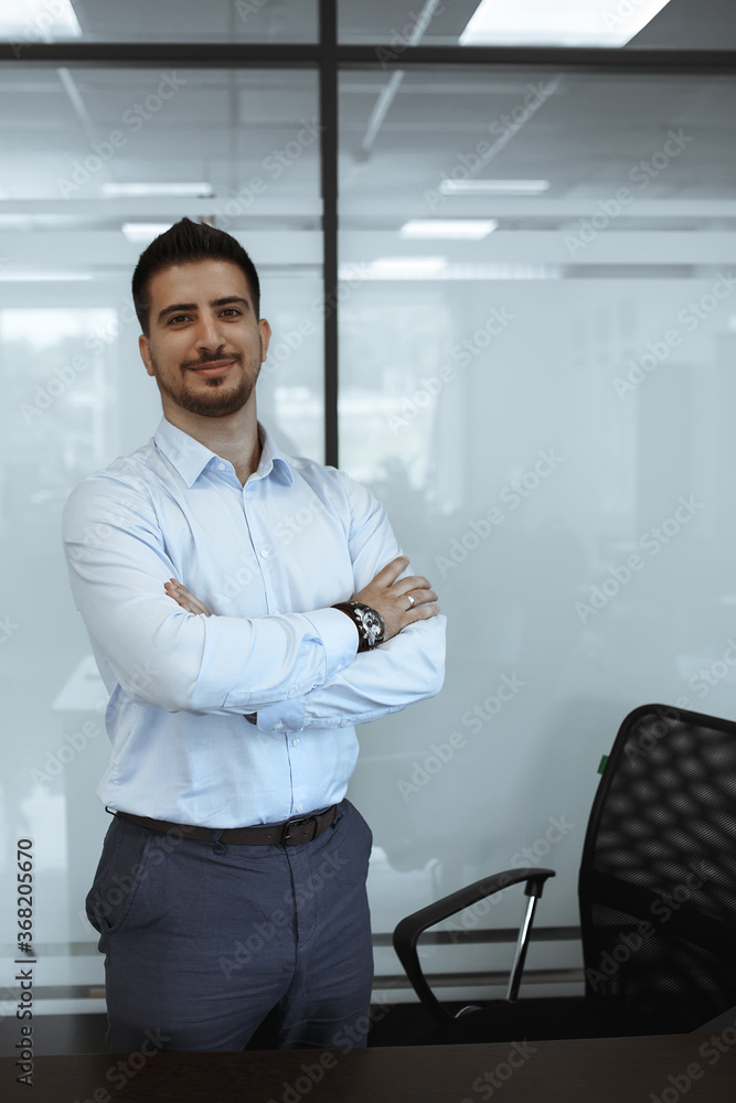 dark-haired man in the office in a blue shirt stands in front of the table with his arms crossed in front of him dark-haired man in the office in a blue shirt stands in front of the table with his arms crossed in front of him