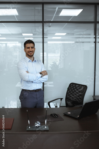 dark-haired man in the office in a blue shirt stands in front of the table with his arms crossed in front of him