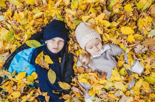 Little brother and sister lie on their backs in yellow leaves. Happy children in autumn park lying on leaves.