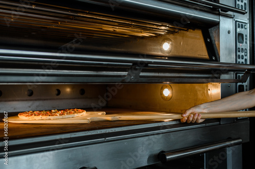 Close up baker is holding a wood peel with fresh pizza in an oven at a baking manufacture factory.