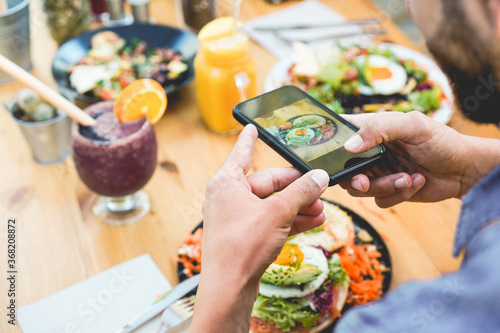 Influencer man eating brunch while making video and photos of dish with mobile phone in trendy bar restaurant - Healthy lifestyle, technology and food trends concept - Focus on man hand