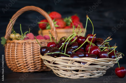 a full plate of ripe cherries with a basket and Cup full of berries in the background on a wooden surface