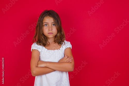 Little caucasian girl with blue eyes wearing white dress standing over isolated red background serious face with crossed arms looking at the camera. Positive person.