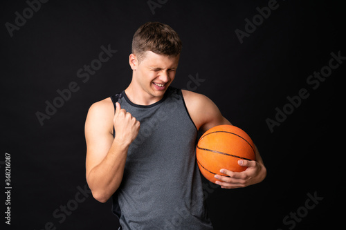 Handsome young man playing basketball over isolated black wall