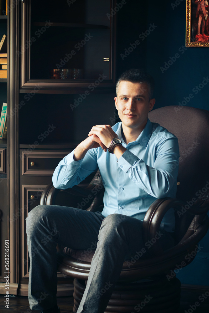 man in a blue shirt smiling sitting in an armchair putting his hands on the armrests and clasping his hands in the lock man in a blue shirt smiling sitting in an armchair putting his hands on the armrests and clasping his hands in the lock