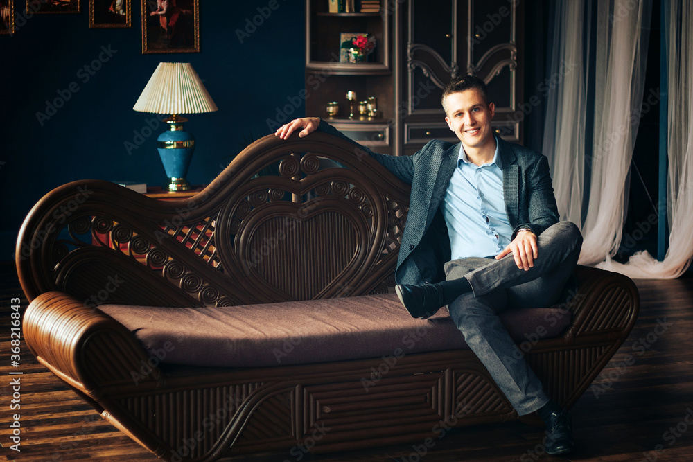 A man in a blue shirt and jacket sits on the couch with one foot on the other.