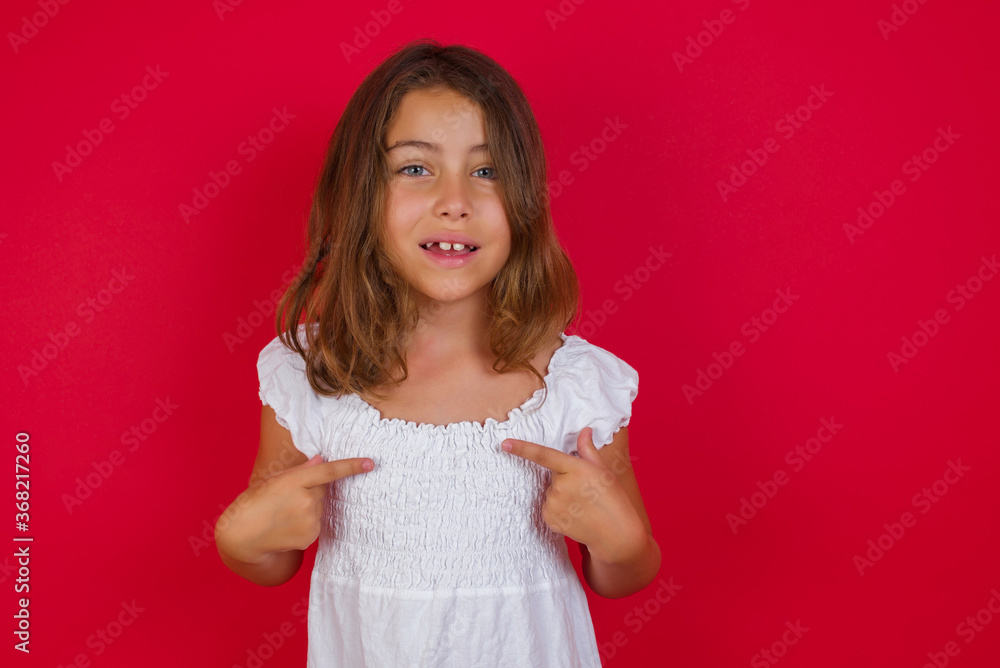 Cheerful stylish Little caucasian girl with blue eyes wearing white dress standing over isolated red wall points at his body, being in good mood after going shopping and making successful purchases Cheerful stylish Little caucasian girl with blue eyes wearing white dress standing over isolated red wall points at his body, being in good mood after going shopping and making successful purchases