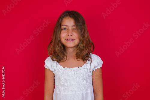Photography Positive coquettish Little caucasian girl with blue eyes wearing white dress  smiling happily, blinking at camera in a playful manner, flirting with you