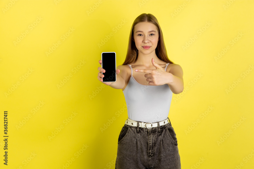 Naklejka premium Pointing on phone's screen. Caucasian young girl's portrait isolated on yellow studio background. Beautiful female blonde model. Concept of human emotions, facial expression, sales, ad, youth culture