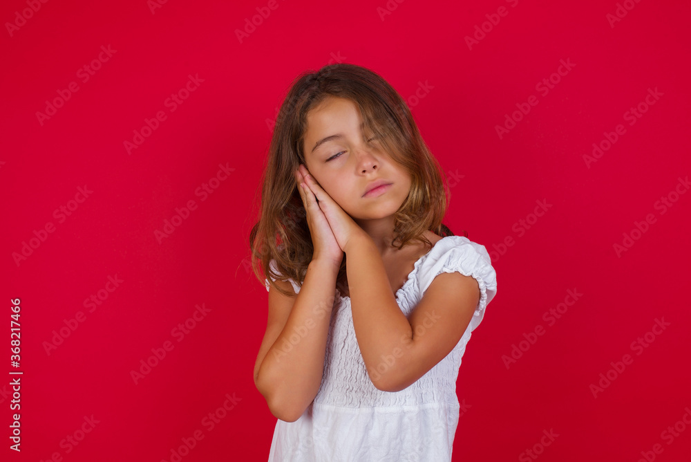 Little caucasian girl with blue eyes wearing white dress standing over isolated red background sleeping tired dreaming and posing with hands together while smiling with closed eyes. Little caucasian girl with blue eyes wearing white dress standing over isolated red background sleeping tired dreaming and posing with hands together while smiling with closed eyes.