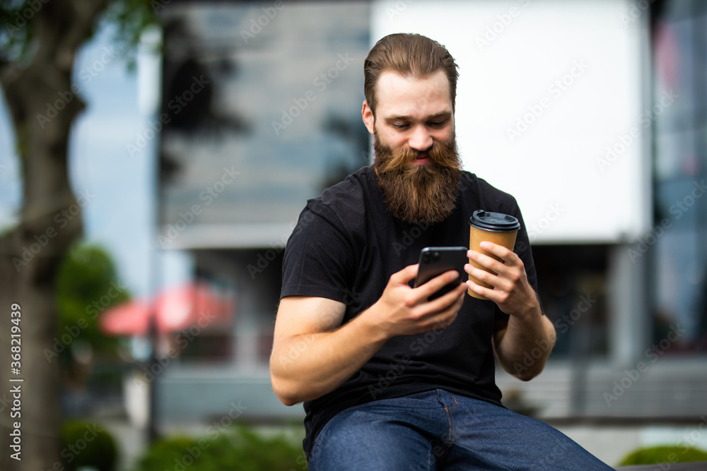 Handsome young brutal bearded man sitting on bench and checking his smart phone outdoors Handsome young brutal bearded man sitting on bench and checking his smart phone outdoors