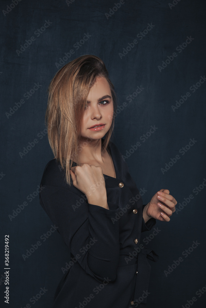 blonde girl with a short haircut on a dark background in a dark blue office dress is half-sided, with her hand on her shoulder blonde girl with a short haircut on a dark background in a dark blue office dress is half-sided, with her hand on her shoulder