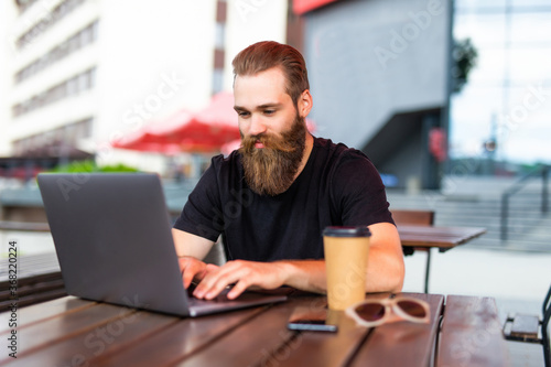 Handsome bearded man using a laptop and taking notes while sitting in cafe in the city center