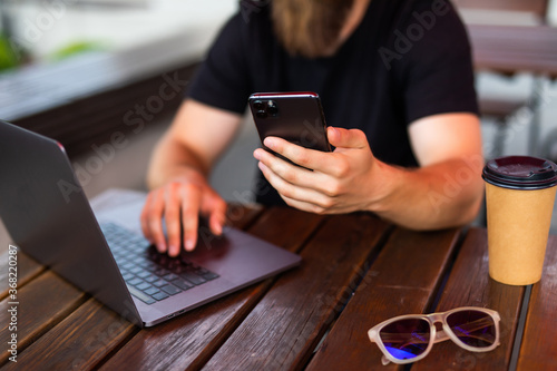 Smiling bearded young man using mobile phone while sitting at table near laptop in street cafe