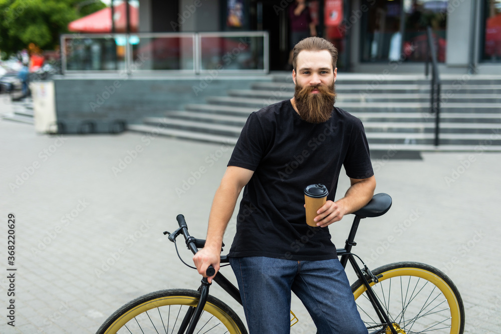 Coffee to go. Side view of young bearded man drinking coffee while sitting on his bicycle outdoors Coffee to go. Side view of young bearded man drinking coffee while sitting on his bicycle outdoors
