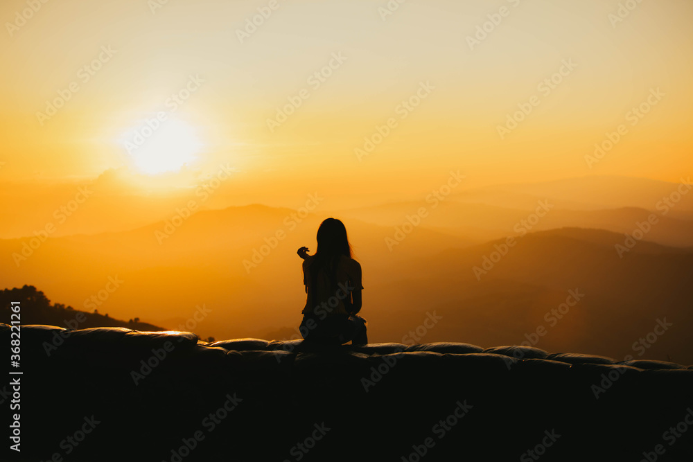 Silhouette of a girl sitting on a cliff side looking at the sunset.  Beautiful sunset background.