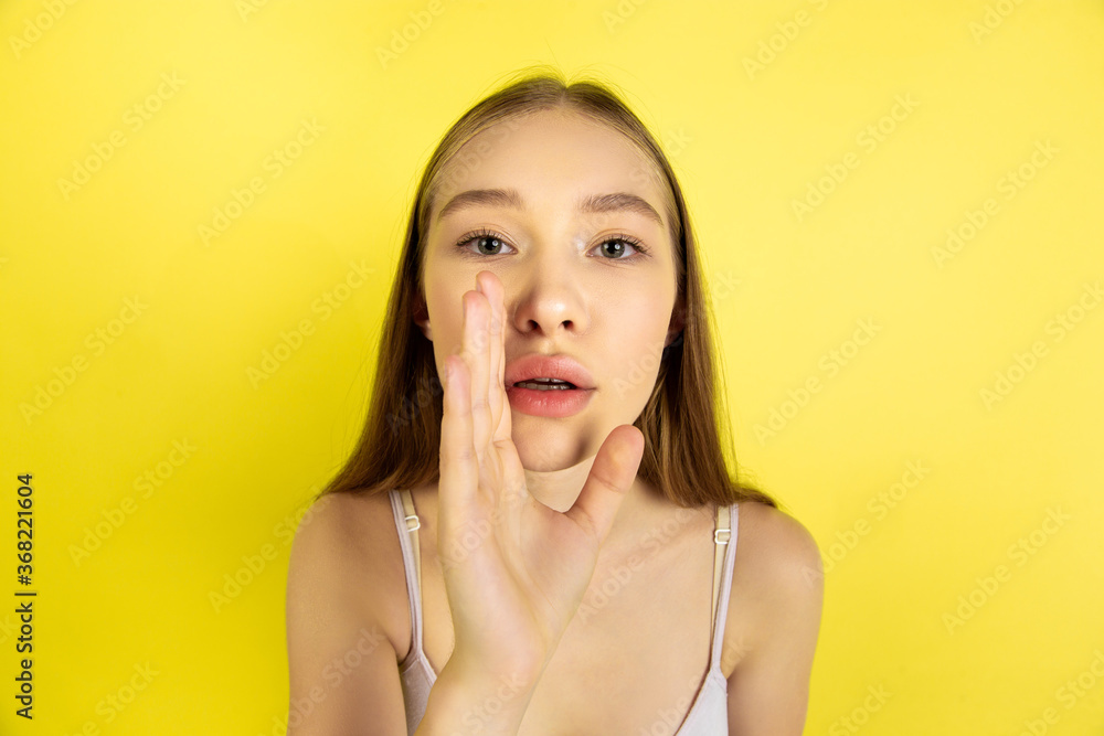 Whispering a secret. Caucasian young girl's portrait isolated on yellow studio background. Beautiful female blonde model. Concept of human emotions, facial expression, sales, ad, youth culture Whispering a secret. Caucasian young girl's portrait isolated on yellow studio background. Beautiful female blonde model. Concept of human emotions, facial expression, sales, ad, youth culture