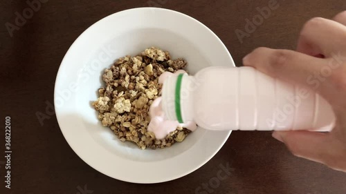 Dry muesli in a white plate on a brown wooden table. Kefir is poured into muesli. A woman's hand pours dry muesli with yogurt and mixes them with a silver spoon