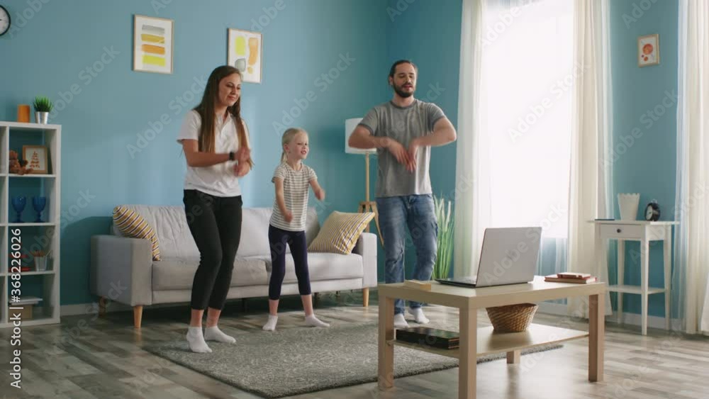 Young family, mother, father, and small girl, are dancing together synchronously, have active morning, practice their favorite dance using laptop, repeat movements after online coach, Slow motion. Young family, mother, father, and small girl, are dancing together synchronously, have active morning, practice their favorite dance using laptop, repeat movements after online coach, Slow motion.
