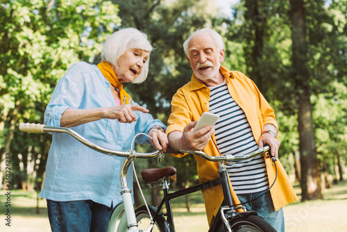 Senior woman pointing at smartphone beside husband and bicycles in summer park