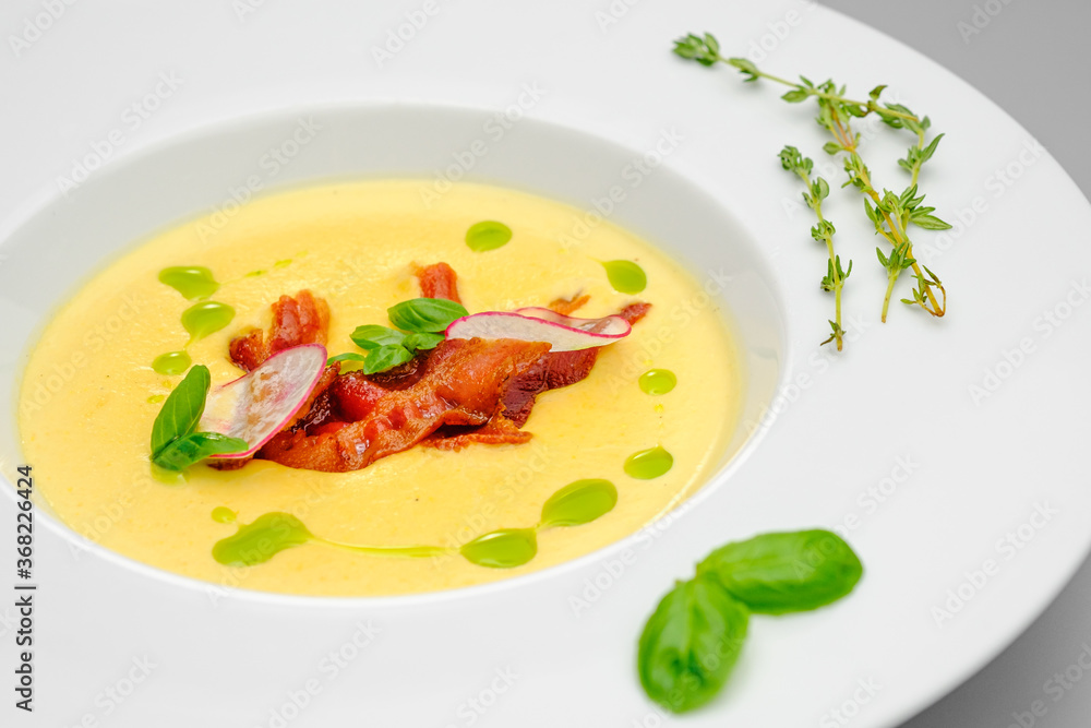 Corn cream soup with smoked meat in a white plate decorated with basil, on a gray background. The first course on the restaurant menu. Corn cream soup with smoked meat in a white plate decorated with basil, on a gray background. The first course on the restaurant menu.