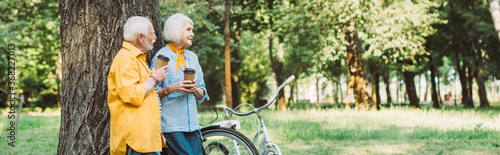 Panoramic shot of smiling senior woman holding disposable cup beside husband and bicycles in park