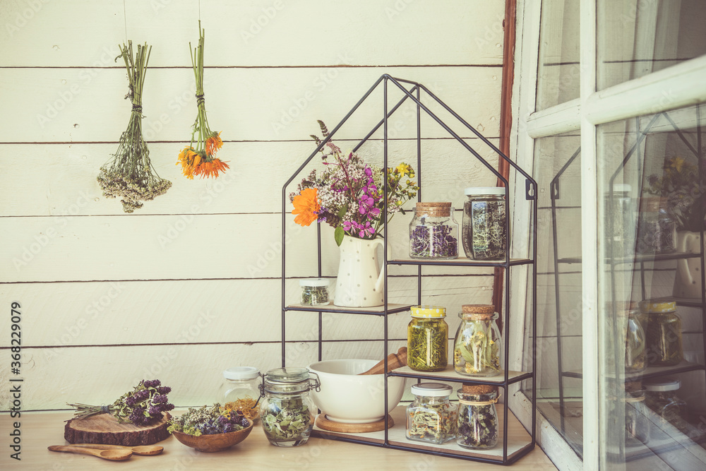 Collection of various herbal medicinal plants dry in glass jars and drying on wall. Modern shelf with jars mortar and pestle and bouquet of herbal wildflowers in ceramic jug. Retro vintage filter.