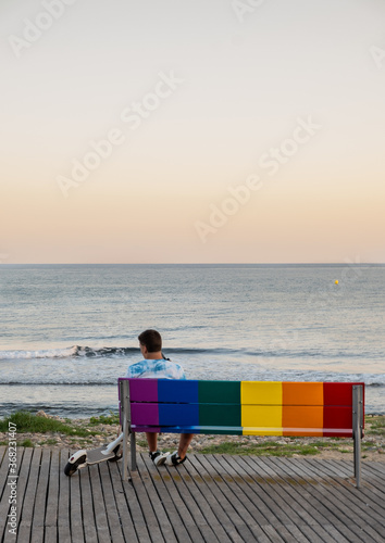 Boy sitting with his scooter on a bench painted in the colors of gay pride overlooking the sea. © daniel_sanfe