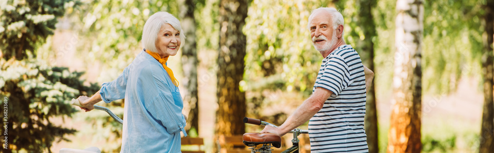 Panoramic crop of senior couple smiling at camera near bicycles in park during summer Panoramic crop of senior couple smiling at camera near bicycles in park during summer