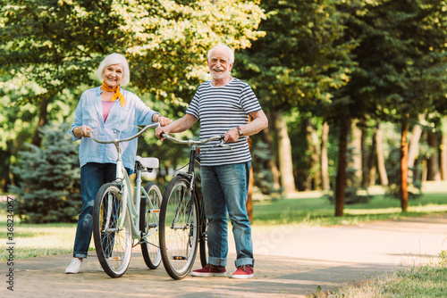 Elderly couple smiling at camera while walking with bicycles on walkway in park