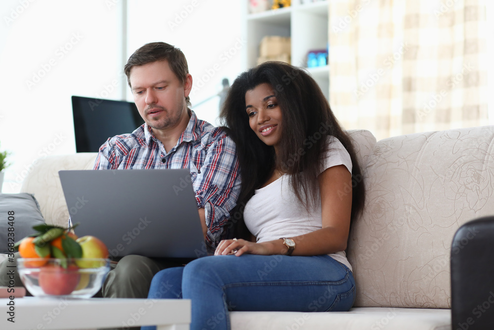 Married couple sitting at home on the couch and looking at the laptop. Young woman enjoys shopping online during a pandemic