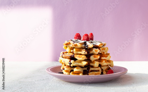Stack of Viennese waffles with raspberries drizzled with chocolate topping in a pink plate. Summer breakfast.