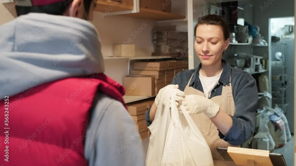 Handheld shot of cheerful female shop employee carrying plastic bags with food and handing them to male client in face mask and gloves Handheld shot of cheerful female shop employee carrying plastic bags with food and handing them to male client in face mask and gloves
