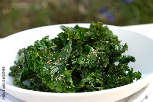 Crunchy baked curly kale chips with sesame seed on white plate. 