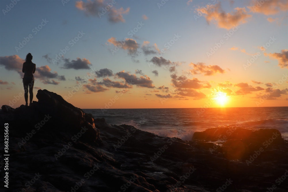 Silhouette of a woman standing on the peak of a big rock on shore watching the sunset 