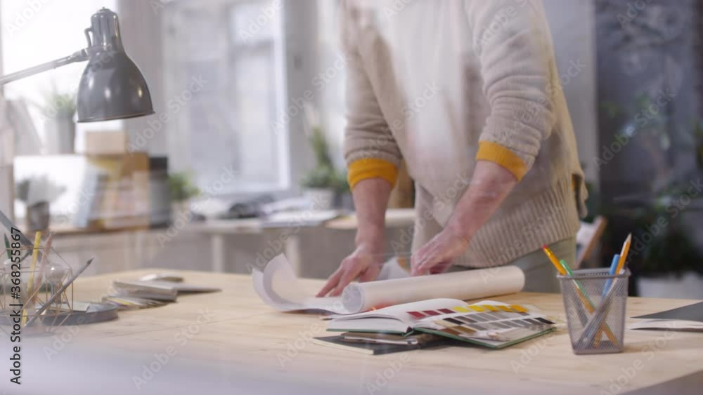 Handheld tracking of middle-aged man in glasses and yellow beanie hat putting blueprint on desk and studying it while working on project in office Handheld tracking of middle-aged man in glasses and yellow beanie hat putting blueprint on desk and studying it while working on project in office