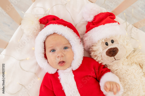 Christmas photo of a baby in a Santa costume lying in a crib at home with a toy in a Santa Claus hat, top view, happy new year