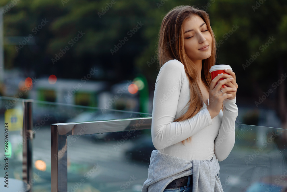 A beautiful young woman with a disposable cup of coffee, enjoy coffee  with closed eyes against the background of the urban city. Urban scene, city life