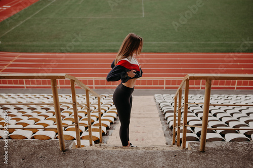 Smiling girl stands at the city stadium, lifts up a sports jacket, and shows her muscles. Mativation concept. Lady with six pack. Progress result after diet and exercise. Image with copy space. photo